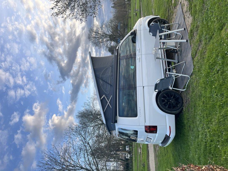 VW California Coast T6.1 mit ausgefahrener Markise, Tisch und Stühlen auf einer Wiese
