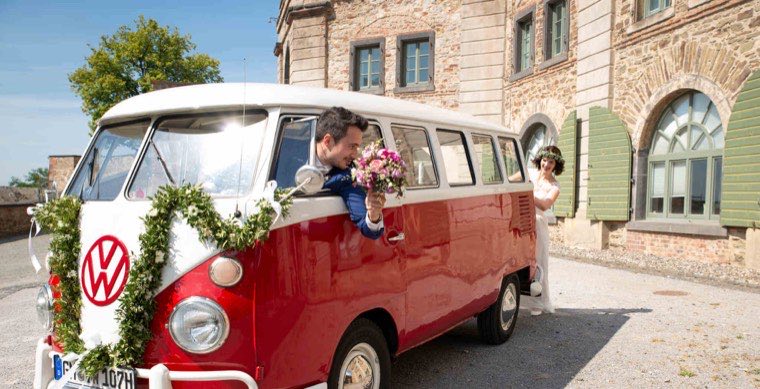 Bräutigam reicht der Braut den Brautstrauß durch das offene Fenster des rot-weißen VW T1 Bulli