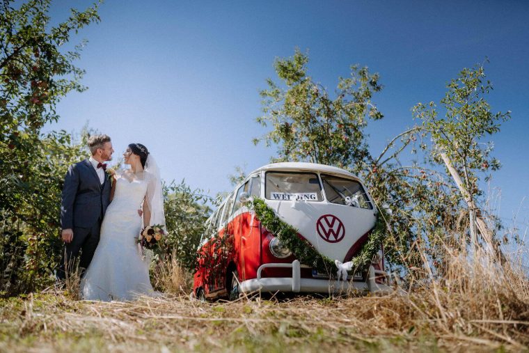 Brautpaar zwischen Apfelbäumen, der rote VW T1 Bulli am Bildrand