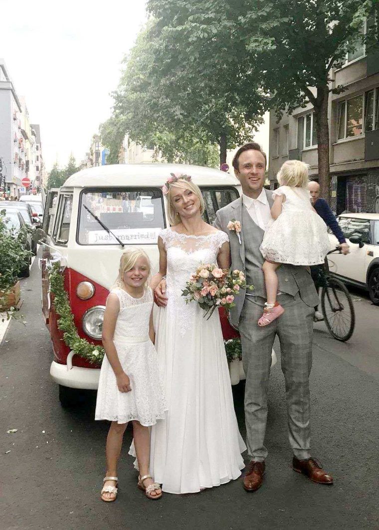 Brautpaar posiert mit zwei Kindern vor dem mit „just married“ beschrifteten Bulli in der Stadt