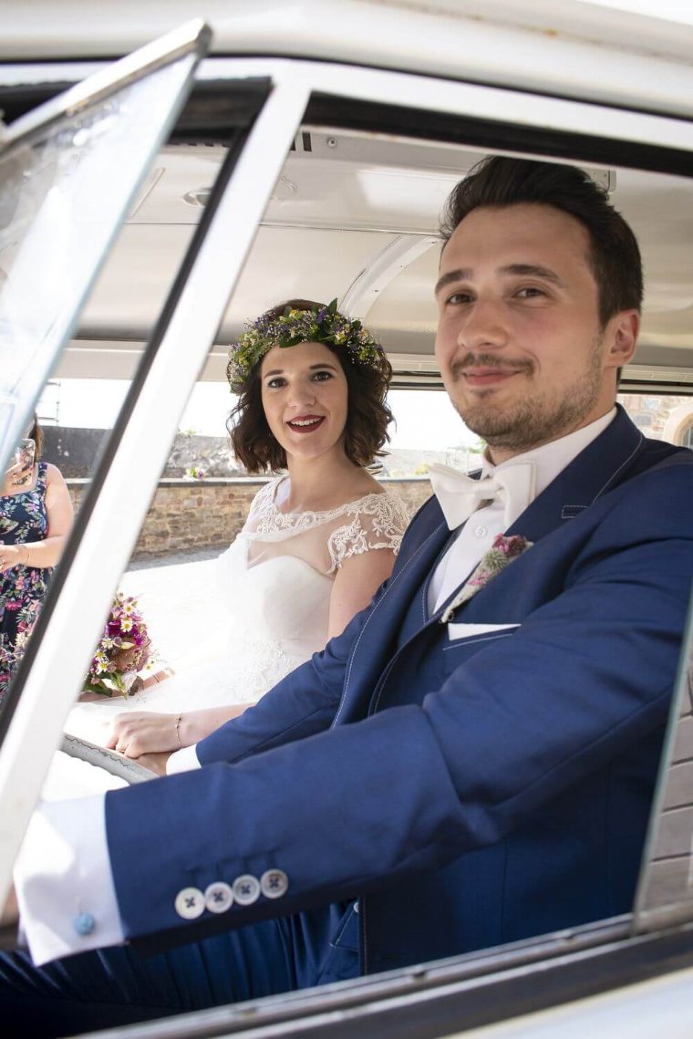 Braut mit Blumenkranz und Bräutigam sitzen nebeneinander im Bulli und blicken aus dem Fenster