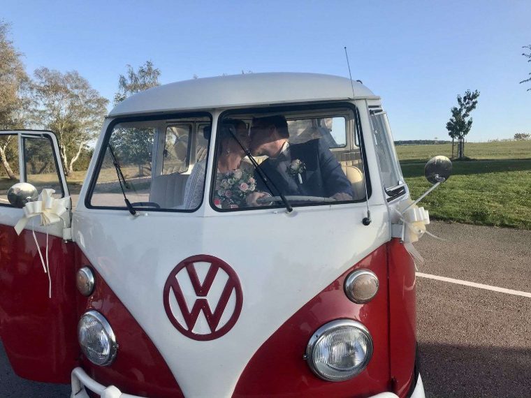 Braut und Bräutigam küssen sich durch das offene Fahrerfenster, weiße Schleifen an den Außenspiegeln