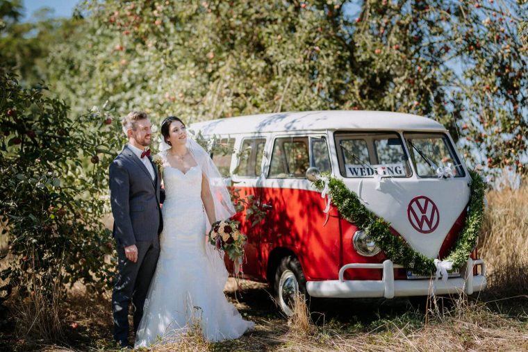 Brautpaar steht in einem Apfelgarten neben dem girlandengeschmückten Bulli, im Fenster ein „WEDDING“-Schild