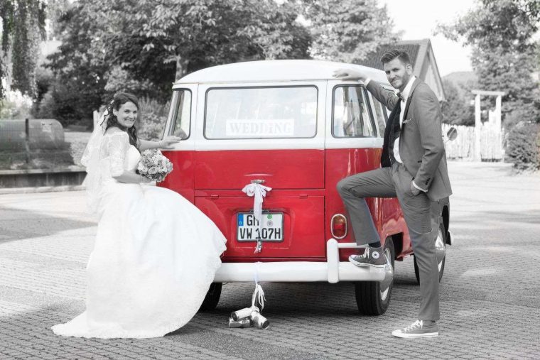 Selektiv coloriertes Foto: Braut und Bräutigam posieren am roten Bulli, im Fenster ein „WEDDING“-Schild, Dosen hängen an der Stoßstange