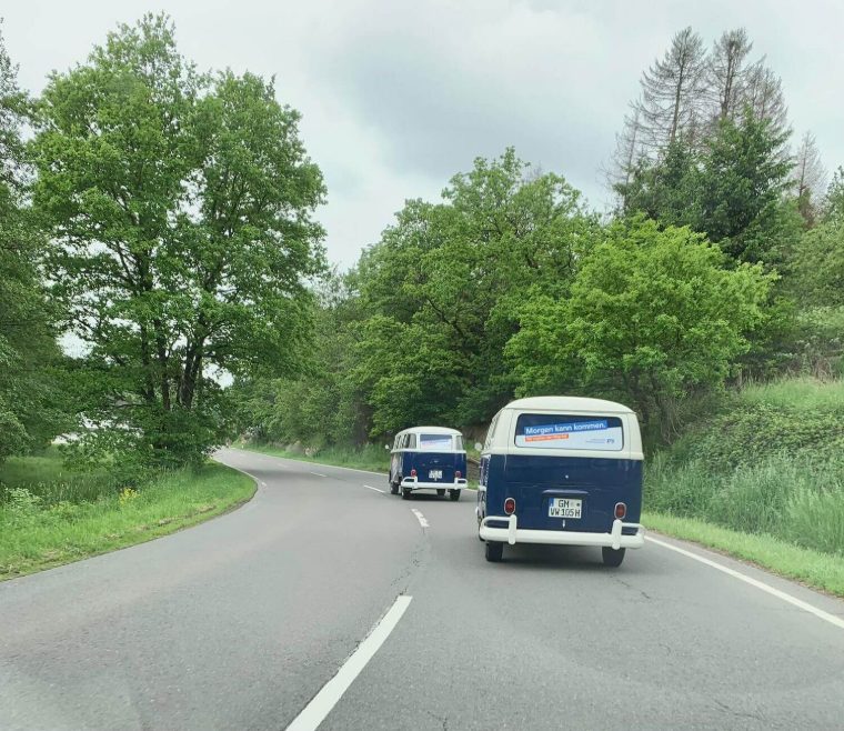 Zwei Bullis fahren hintereinander auf einer Landstraße durchs Grüne