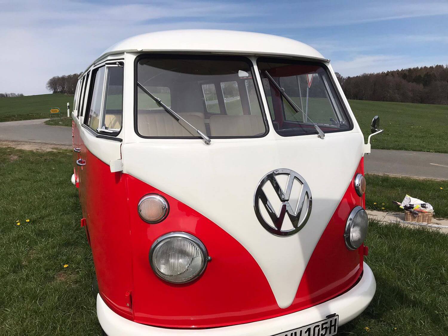 Rot-weißer VW T1 Bulli Baujahr 1964 auf einer grünen Wiese am Straßenrand, im Hintergrund sanfte Hügel