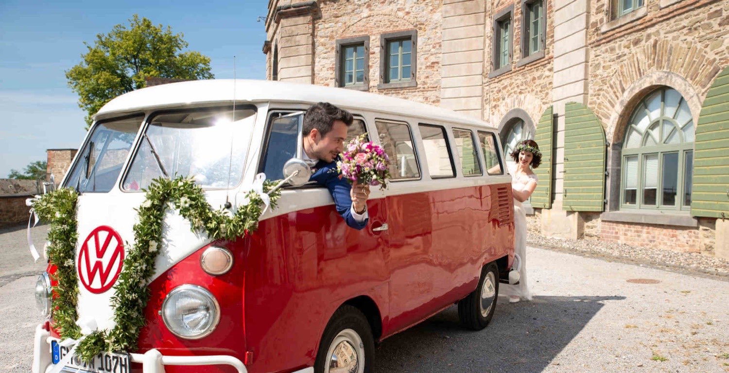 Bräutigam reicht der Braut den Brautstrauß durch das offene Fenster des rot-weißen VW T1 Bulli