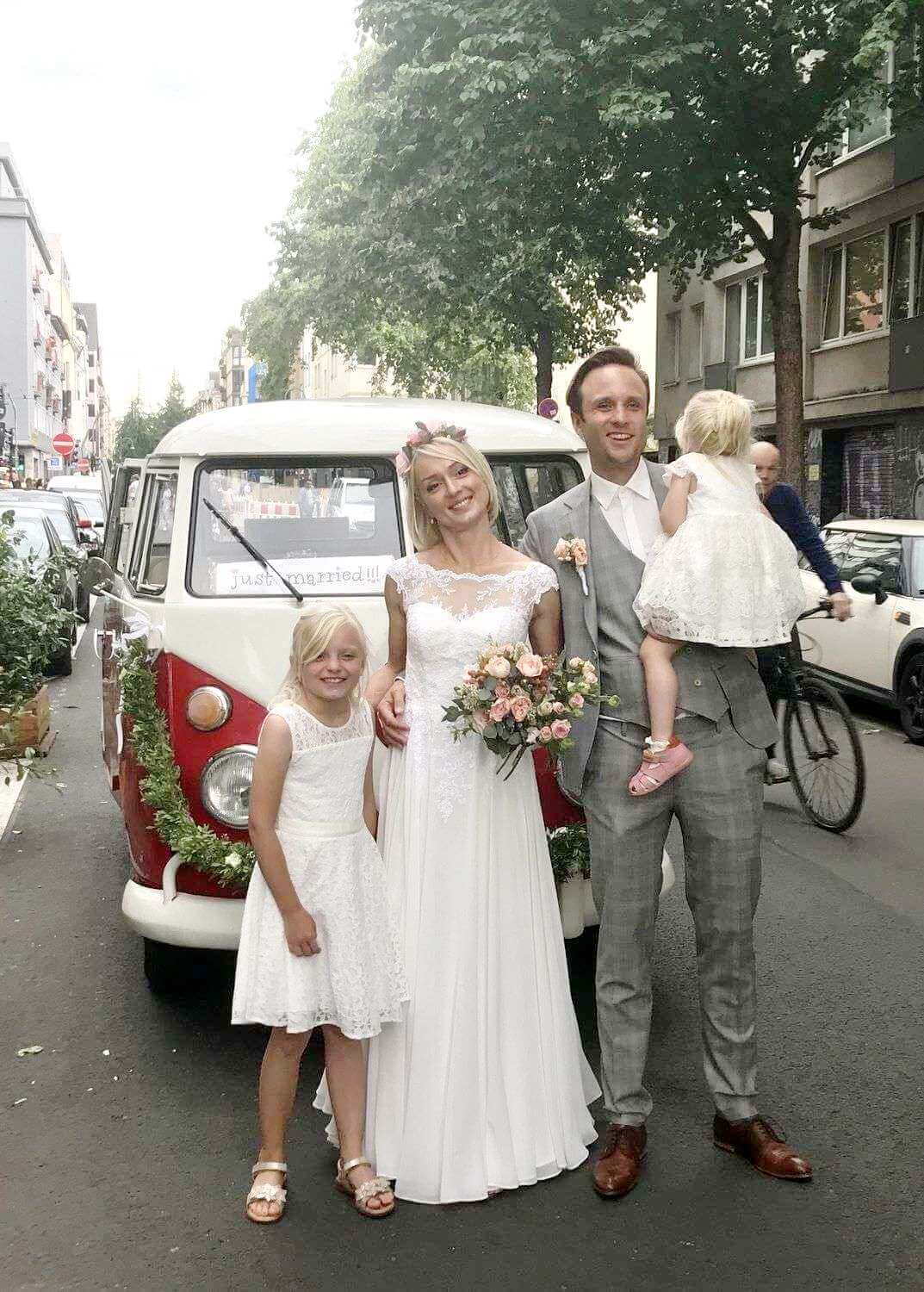 Brautpaar posiert mit zwei Kindern vor dem mit „just married“ beschrifteten VW T1 Bulli mitten in der Stadt