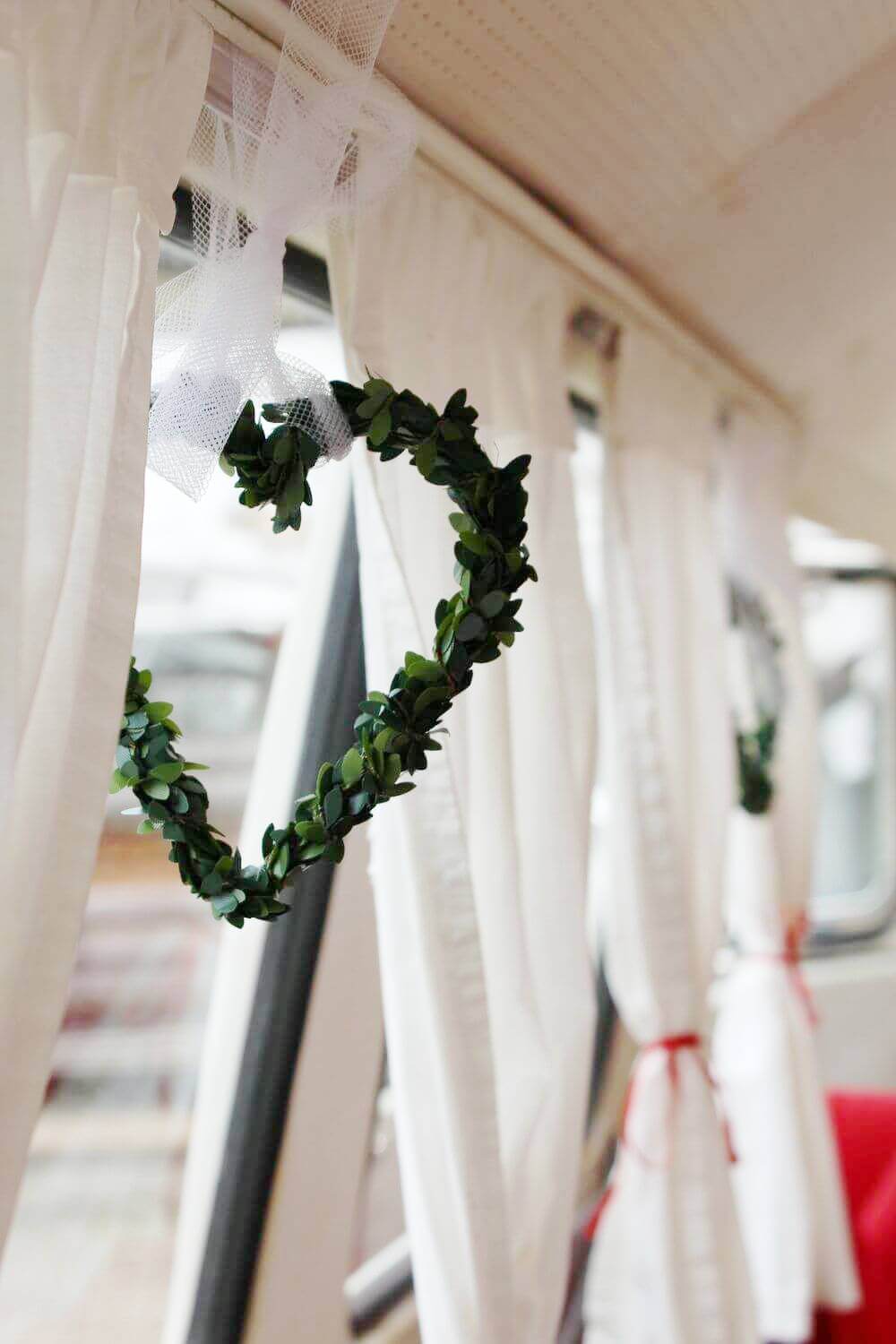 Herz aus Buchsbaumzweigen hängt an der weißen Fenstergardine im Innenraum des VW T1 Bulli