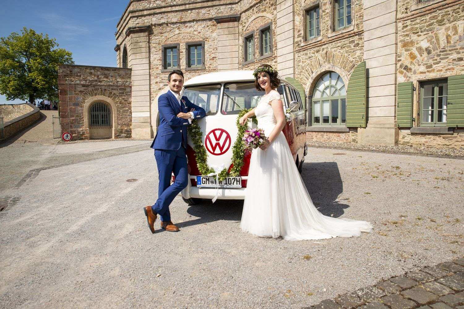 Brautpaar vor dem rot-weißen VW T1 Bulli auf einem historischen Hof
