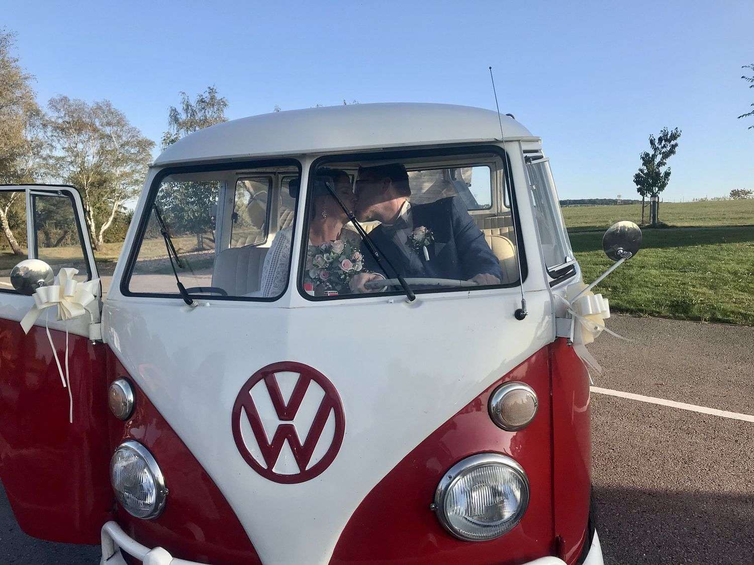Braut und Bräutigam küssen sich durch das offene Fahrerfenster des VW T1 Bulli, weiße Schleifen an den Außenspiegeln