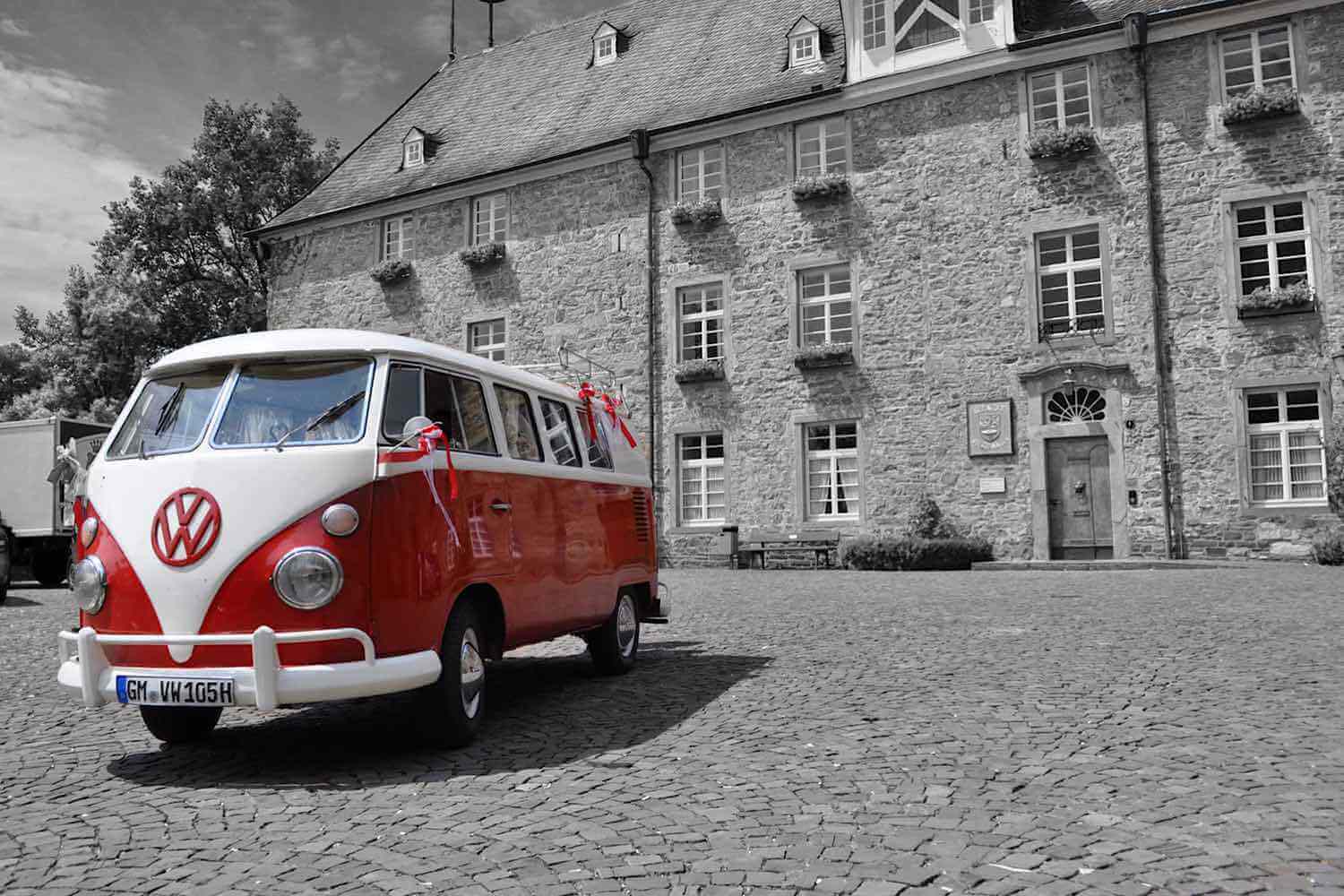 Rot-weißer VW T1 Bulli mit Dekoschleifen vor einem historischen Schlossgebäude, Schwarz-Weiß-Aufnahme mit Farbakzent