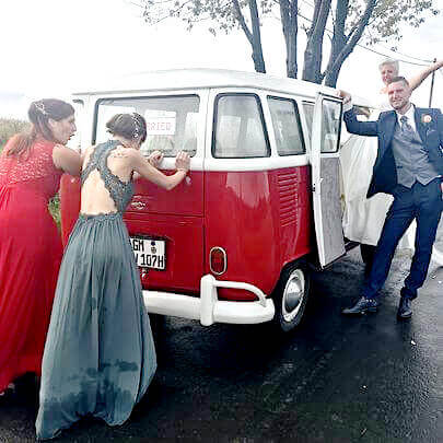 Brautpaar mit VW Bus Oldtimer auf der Strasse und zwei elegant gekleidete Gäste schieben.